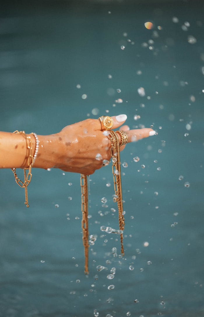 A graceful hand wearing gold bracelets and rings interacts with splashing water.